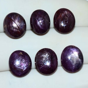 May include: Six oval-shaped, deep purple cabochon gemstones arranged in two rows. Each stone features a smooth, polished surface with visible internal inclusions and a distinct star-like light reflection pattern across the dark, translucent surfaces.