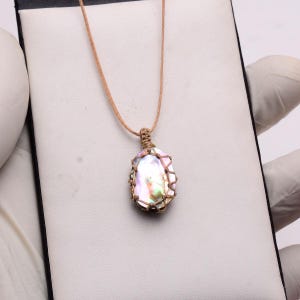 Könnte beinhalten: Eine perlmuttfarbene Anhängerkette mit einer neutralfarbenen Kordel. Der Anhänger hat eine ovale Form und ist in einem gewebten, hellbraunen Material eingeschlossen. Der schillernde Stein reflektiert Farben von Rosa, Grün und Gelb.
