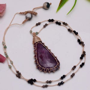 Può includere: Una collana con una grande gemma sfaccettata a forma di goccia di colore viola intenso, incastonata in un cordoncino di macramè color cuoio. Il cordoncino è decorato con piccole perline nere e grigie, e la chiusura regolabile include due perline rotonde in pietra grigia e bianca.