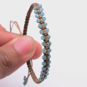 May include: A woven bracelet with light blue gemstone beads and a brown cord. The bracelet has an adjustable sliding knot closure. The small, faceted beads reflect light. The cord is a natural, earthy tone.