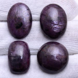 May include: Four polished, oval and round gemstones in shades of purple and burgundy. The gemstones have a smooth, reflective surface with visible internal patterns. The stones are displayed on a white, textured fabric.