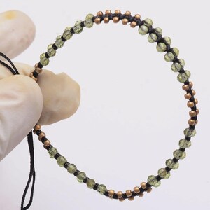 May include: A circular bracelet constructed from black cord featuring alternating sections of small, metallic copper-colored beads and faceted, translucent olive-green beads. The design is arranged in a repeating pattern along the entire length of the cord.