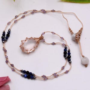 May include: A necklace featuring a clear, faceted, oval-shaped pendant encased in tan cord macrame. The cord is adorned with small, light purple, dark blue, and black beads, and includes two white spherical beads at the adjustable sliding closure.
