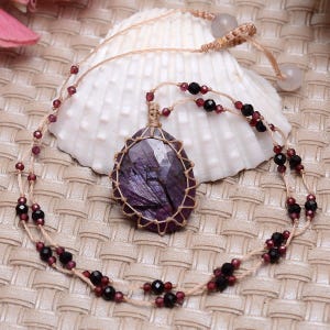 May include: A necklace featuring a faceted, teardrop-shaped purple gemstone encased in a tan macrame cord netting. The cord is accented with small, dark red and black faceted beads, and the necklace includes two light-coloured round beads at the closure.