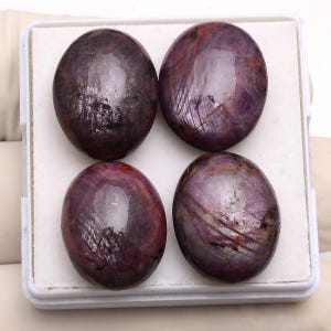 May include: Four oval-shaped, polished gemstones in a deep burgundy hue with a glossy finish. The stones, set in a white tray, appear to be ruby cabochons. The gems display internal striations and colour variations.