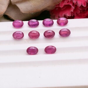 Puede incluir: Diez rubíes sueltos de corte ovalado con un tono magenta rosado vibrante y superficies facetadas. Estas piedras preciosas están dispuestas en tres filas sobre una bandeja de exhibición blanca escalonada, mostrando su calidad translúcida.