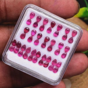 Puede incluir: Un estuche de presentación cuadrado de plástico transparente que contiene una colección de pequeños rubíes facetados en forma de pera. Las vibrantes gemas de color rojo rosado intenso están dispuestas en filas sobre espuma blanca.
