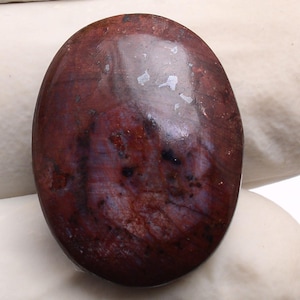 Könnte beinhalten: Oval geschliffener Edelstein in tiefen Burgunder- und Lilatönen. Der Stein hat eine glatte, glänzende Oberfläche mit natürlichen Adern und Mustern. Wahrscheinlich für Schmuck oder Dekorationszwecke verwendet.