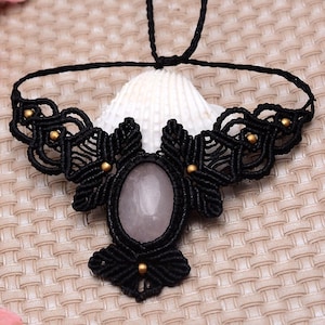Op de afbeelding: Een zwarte macramé ketting met een ovaalvormige, lichtroze rozenkwarts edelsteen in het midden. Het ingewikkelde ontwerp van geknoopte koorden bevat kleine, ronde, goudkleurige metalen kralen die in het patroon zijn verwerkt.