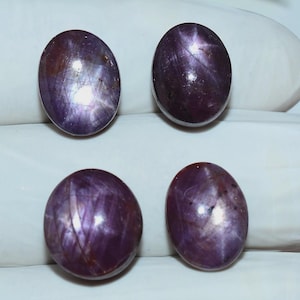 May include: Four oval-shaped, polished gemstones with a deep purple hue. Each stone displays a star-like effect, with lines radiating from a central point. The gems are smooth and reflect light, suggesting a high-quality finish.