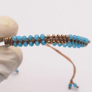 Könnte beinhalten: Ein handgefertigtes Armband mit kleinen, runden, hellblauen Perlen auf einer braunen Kordel. Das Armband hat ein gewebtes Design mit goldfarbenen Metallakzenten. Die Kordel geht in einen Kordelverschluss über.