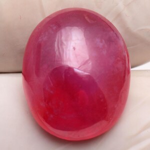May include: A smooth, oval-shaped gemstone with a polished surface and a vibrant, translucent deep pink color. The stone features subtle internal cloud-like inclusions and a high-gloss finish that reflects light across its rounded, dome-like top.