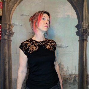 Może przedstawiać: Czarny t-shirt z dekoracyjnym, ażurowym karczkiem z koronki z motywem serca. Koronka jest barwiona na złoty kolor. Modelka ma na sobie dżinsy w ciemnym odcieniu. Tło to malowidło ścienne z elementami architektonicznymi i latającymi spodkami.