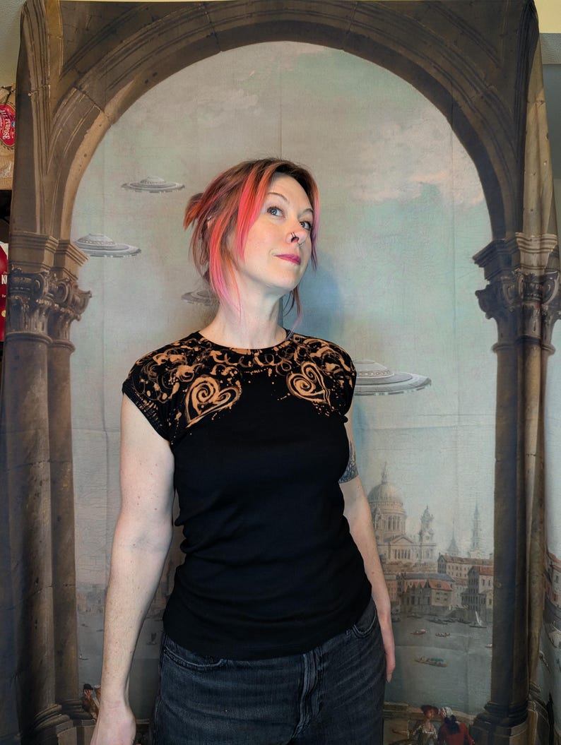 Może przedstawiać: Czarny t-shirt z dekoracyjnym, ażurowym karczkiem z koronki z motywem serca. Koronka jest barwiona na złoty kolor. Modelka ma na sobie dżinsy w ciemnym odcieniu. Tło to malowidło ścienne z elementami architektonicznymi i latającymi spodkami.