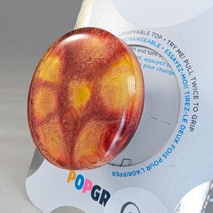 May include: A round, red, yellow, and orange resin button with a swirling pattern. The button is on a white background with the text "ESSAYEZ-MOI! TIREZ-LE DEUX FOIS POUR L'AGRIPPER" and "SWAPPABLE TOP TRY ME! PULL TWICE TO GRIP CHANGEABLE" printed on it.