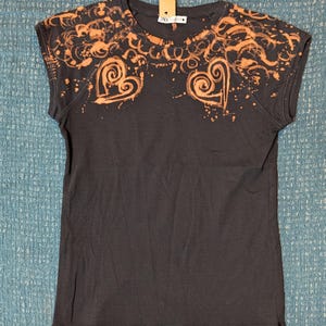 Może przedstawiać: Ciemnoszara koszulka z dekoltem w serek i kr&oacute;tkimi rękawami. Koszulka ma pomarańczowy wz&oacute;r z wir&oacute;w i kształt&oacute;w serca na ramionach i klatce piersiowej, z efektem rozprysku. Etykieta jest przymocowana do dekoltu.