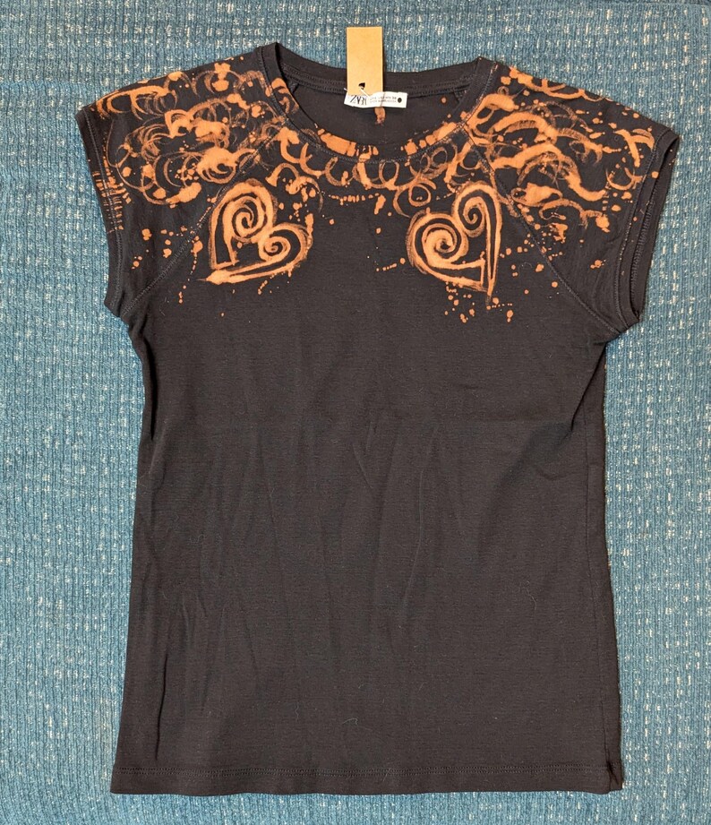 Może przedstawiać: Ciemnoszara koszulka z dekoltem w serek i kr&oacute;tkimi rękawami. Koszulka ma pomarańczowy wz&oacute;r z wir&oacute;w i kształt&oacute;w serca na ramionach i klatce piersiowej, z efektem rozprysku. Etykieta jest przymocowana do dekoltu.