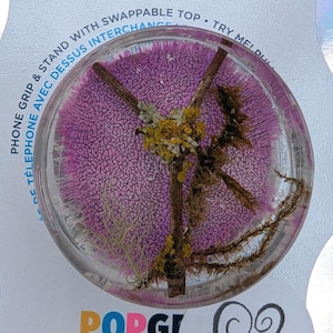May include: A clear phone grip with a pink, textured base and a variety of dried plants and flowers embedded inside. The text "PHONE GRIP & STAND WITH SWAPPABLE TOP" is printed on the white background.