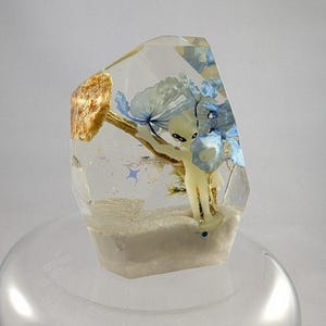 May include: A clear resin sculpture with a geometric shape. Inside, a small, pale figure stands amidst blue flowers and gold accents. The base is a white, sandy texture. The sculpture is on a clear, rounded stand.