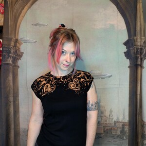 Może przedstawiać: Czarny t-shirt z kr&oacute;tkim rękawem z motywem serca na ramionach. Wz&oacute;r serca jest w kolorze jasno pomarańczowym. Osoba na zdjęciu ma na sobie ciemnoszare dżinsy. Tło to malowidło ścienne z kolumnami i pejzażem miejskim.