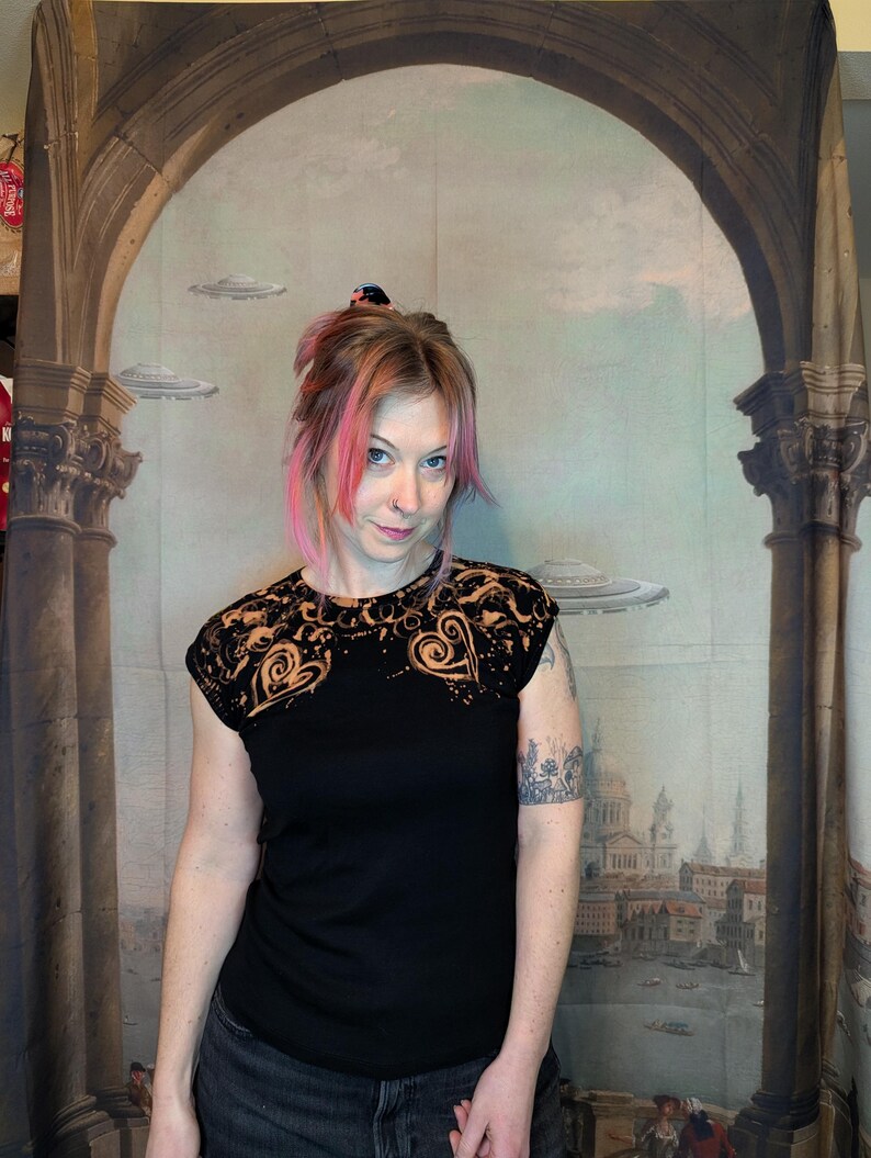 Może przedstawiać: Czarny t-shirt z kr&oacute;tkim rękawem z motywem serca na ramionach. Wz&oacute;r serca jest w kolorze jasno pomarańczowym. Osoba na zdjęciu ma na sobie ciemnoszare dżinsy. Tło to malowidło ścienne z kolumnami i pejzażem miejskim.