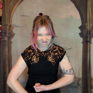 Może przedstawiać: Czarny t-shirt z wybielonym wzorem na ramionach i rękawach. Koszulka ma okrągły dekolt i kr&oacute;tkie rękawy. Tło to malowidło ścienne z łukami i latającymi spodkami.