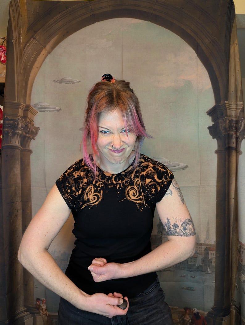 Może przedstawiać: Czarny t-shirt z wybielonym wzorem na ramionach i rękawach. Koszulka ma okrągły dekolt i kr&oacute;tkie rękawy. Tło to malowidło ścienne z łukami i latającymi spodkami.