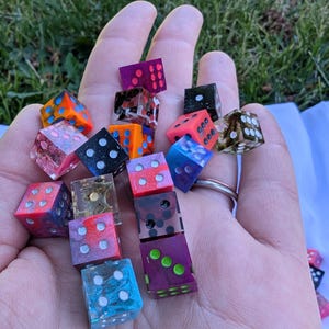 May include: A collection of small, multi-colored resin dice featuring various patterns, including glitter, swirls, and solid colors. Each die has six sides with circular pips in contrasting colors, showcasing a variety of vibrant hues like blue, pink, orange, purple, and black.
