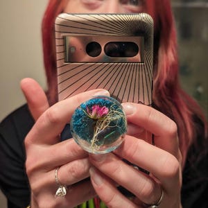 May include: A clear phone grip with a pink flower and blue resin inside. The grip is attached to a black and white striped phone case.