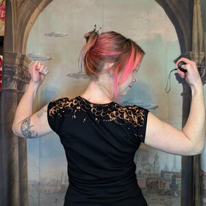 Może przedstawiać: Czarny t-shirt z wybielonym wzorem na ramionach. Osoba prezentuje zgięte ramiona, pokazując tył koszulki. Tło to malowidło ścienne z łukami, latającymi spodkami i krajobrazem miejskim.