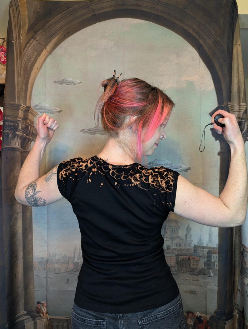 Może przedstawiać: Czarny t-shirt z wybielonym wzorem na ramionach. Osoba prezentuje zgięte ramiona, pokazując tył koszulki. Tło to malowidło ścienne z łukami, latającymi spodkami i krajobrazem miejskim.