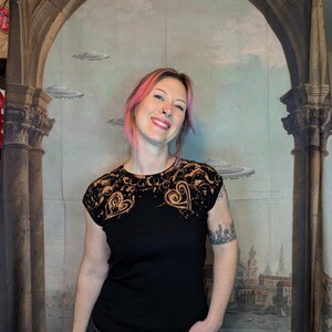 Może przedstawiać: Czarny t-shirt z dekoracyjnym wzorem wok&oacute;ł dekoltu i ramion. Wz&oacute;r przedstawia kształty serc i wiry w kontrastowym kolorze. Koszulka ma kr&oacute;tkie rękawy i okrągły dekolt.