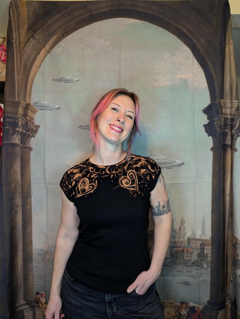 Może przedstawiać: Czarny t-shirt z dekoracyjnym wzorem wok&oacute;ł dekoltu i ramion. Wz&oacute;r przedstawia kształty serc i wiry w kontrastowym kolorze. Koszulka ma kr&oacute;tkie rękawy i okrągły dekolt.