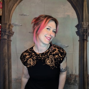 Może przedstawiać: Czarny t-shirt z wybielonym wzorem w kształcie serc i wir&oacute;w. Koszulka ma okrągły dekolt i kr&oacute;tkie rękawy. Model uśmiecha się na tle tła z łukami, kolumnami i latającymi spodkami.