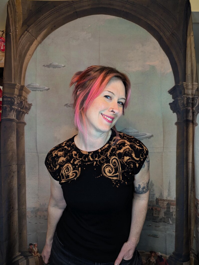 Może przedstawiać: Czarny t-shirt z wybielonym wzorem w kształcie serc i wir&oacute;w. Koszulka ma okrągły dekolt i kr&oacute;tkie rękawy. Model uśmiecha się na tle tła z łukami, kolumnami i latającymi spodkami.