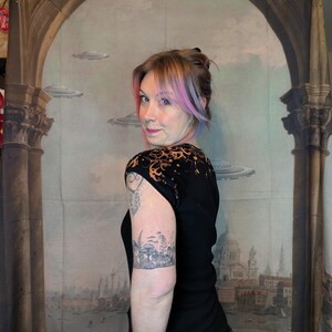 Może przedstawiać: Czarny top z kr&oacute;tkim rękawem z unikalnym wzorem na ramionach. Top jest połączony z ciemnymi dżinsami. Osoba na zdjęciu ma tatuaż na ramieniu. Tło przedstawia mural z elementami architektonicznymi i latającymi spodkami.