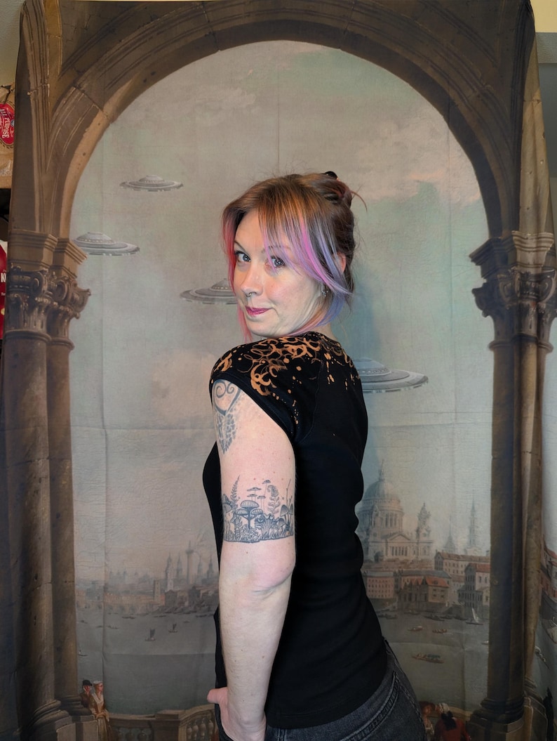 Może przedstawiać: Czarny top z kr&oacute;tkim rękawem z unikalnym wzorem na ramionach. Top jest połączony z ciemnymi dżinsami. Osoba na zdjęciu ma tatuaż na ramieniu. Tło przedstawia mural z elementami architektonicznymi i latającymi spodkami.