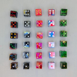 May include: A collection of twenty-five colorful dice with various designs. The dice feature heart, dot, and abstract patterns in a variety of colors, including red, green, blue, and gold. Each die has six sides, with the pips or designs on each side.