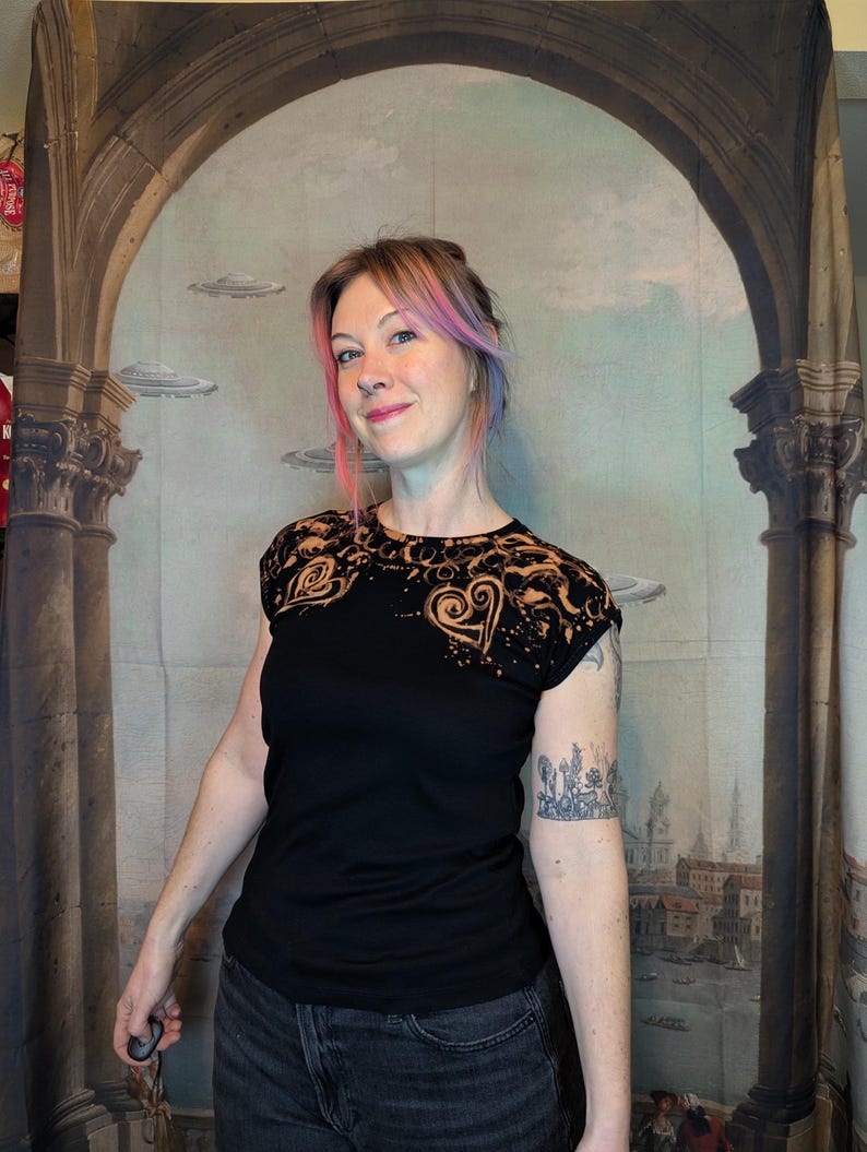 Może przedstawiać: Czarny t-shirt z motywem serca na ramieniu i klatce piersiowej. Wz&oacute;r ma wybielony efekt, tworząc unikalny wz&oacute;r. Modelka ma na sobie ciemne dżinsy i tatuaż na ramieniu.