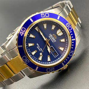 May include: A two-tone wristwatch with a blue face and a rotating blue bezel. The watch has gold-coloured accents and a stainless steel strap. The watch face displays the brand name "Orient" and a date window. The strap is a combination of silver and gold links.