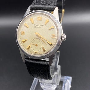 Puede incluir: Un reloj de pulsera SVALAN Sportsman vintage con caja plateada y correa de cuero negro. La esfera del reloj es de color crema claro con marcadores y agujas doradas. El reloj tiene un pequeño dial de segundos y la palabra "WATERPROOF" es visible.