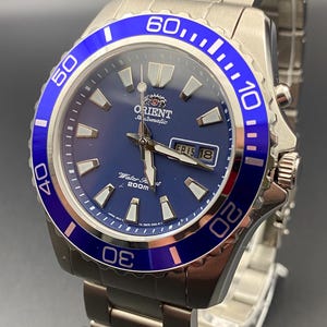 May include: A stainless steel Orient automatic watch with a blue dial and bezel. The watch features a date display and luminous hands and markers. The bezel has white numerals and markers. The watch is on a stainless steel bracelet.