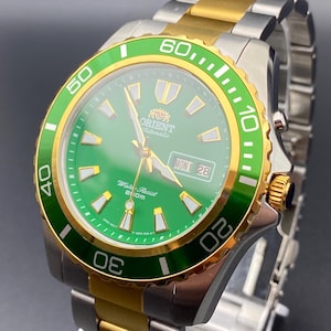 May include: A stainless steel and gold-tone Orient automatic watch with a green dial. The watch features a rotating bezel with white markings, a date display, and the text "Water Resist 200m". The watch band is a combination of silver and gold.