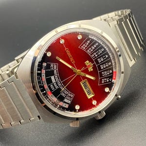 以下が含まれることがあります： 赤い文字盤とカレンダー機能が付いた、シルバーカラーのオリエントの腕時計。ステンレススチール製のバンドと、3時の位置に日付表示があります。文字盤にはゴールドの針とマーカーがあり、6時の位置に曜日表示があります。