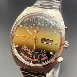 Può includere: Un orologio Orient in acciaio inossidabile con quadrante dorato. L'orologio presenta un display giorno e data, insieme a una funzione calendario. L'orologio ha un cinturino in metallo ed è impostato su lunedì 30.