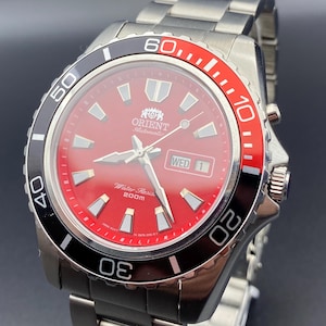 Può includere: Un orologio automatico Orient in acciaio inossidabile con quadrante rosso e lunetta nera e rossa. L'orologio presenta lancette e indici argentati, un display giorno-data e la scritta "Water Resist 200m". Anche il cinturino è in acciaio inossidabile.