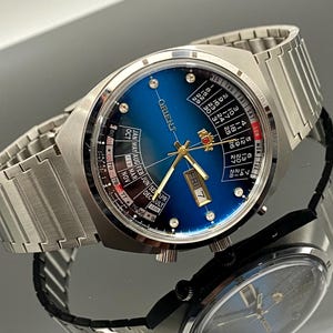 May include: A silver Orient watch with a blue face and calendar features. The watch shows the day, date, and month. It has a silver metal strap and is displayed on a reflective surface.