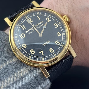 K&ouml;nnte beinhalten: Eine goldfarbene Armbanduhr mit schwarzem Zifferblatt und schwarzem Lederarmband. Das Zifferblatt hat goldene Ziffern und Zeiger mit dem Text "Ulysse Nardin Locle Suisse 1846". Die Uhr befindet sich an einem Handgelenk.