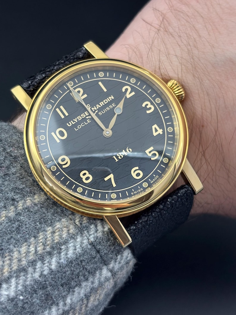 K&ouml;nnte beinhalten: Eine goldfarbene Armbanduhr mit schwarzem Zifferblatt und schwarzem Lederarmband. Das Zifferblatt hat goldene Ziffern und Zeiger mit dem Text "Ulysse Nardin Locle Suisse 1846". Die Uhr befindet sich an einem Handgelenk.