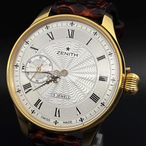 May include: A gold-tone Zenith wristwatch with a silver dial featuring a wavy guilloché pattern, black Roman numerals, and a sub-dial for seconds. The watch includes a dark brown textured leather strap and a gold-tone crown on the right side.